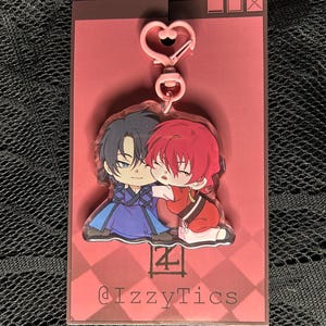 May include: A clear acrylic keychain featuring two anime-style characters embracing. One character wears blue, the other red. The keychain has a pink heart-shaped clasp and is attached to a pink backing card with the text "@IzzyTics".