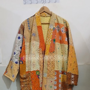 May include: A long, patchwork quilt robe with a V-neck and long sleeves. The robe is made of a variety of fabrics in shades of orange, brown, and yellow. The fabric is quilted with a small, repeating pattern. The robe is hanging on a wooden hanger.