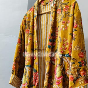 Puede incluir: Una bata de color amarillo mostaza con un estampado floral vibrante en tonos rosas, rojos y verdes. La prenda tiene mangas largas con puños doblados que revelan un forro de rayas verticales blancas y grises, que también recubre el interior.
