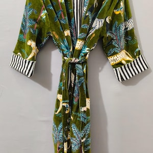 May include: Green velvet robe with a jungle print featuring leopards, palm trees, and other foliage. The robe has a striped black and white interior and cuffs, and a matching belt. The robe is hanging on a wooden hanger.