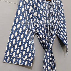 Puede incluir: Bata azul con un estampado de peces blancos, colgada de una percha de madera. La bata tiene mangas largas y un cierre de lazo. El patrón de peces se repite en toda la bata.
