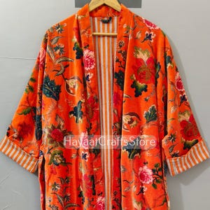 May include: An orange floral print robe with long sleeves. The robe features a vibrant pattern of red, pink, and green flowers and leaves. The interior and sleeve cuffs have a white and orange striped design. The text "Hayaat Crafts Store" is visible.