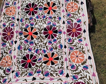 May include: A white textile featuring intricate hand-embroidered floral patterns in vibrant shades of magenta, orange, blue, and black. The design includes large circular flower motifs surrounded by smaller buds and winding green vines.