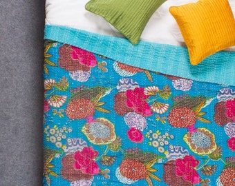 May include: A turquoise blue kantha quilt with a floral pattern in shades of pink, brown, and white. The quilt is folded and displayed on a white bed with two decorative pillows, one green and one yellow.