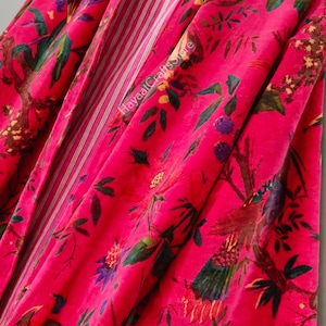 May include: A vibrant pink floral print robe with a striped interior lining. The robe features a botanical design with green leaves, purple flowers, and brown branches. The robe is hanging on a wooden hanger.