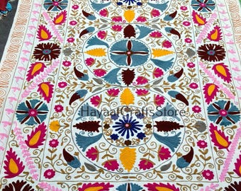 May include: White textile with intricate embroidery in a symmetrical floral pattern. The design features blue, pink, yellow, and brown flowers and leaves. The text "Hayaal Crafts Store" is visible. The border has a repeating floral pattern.