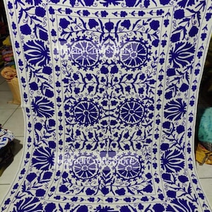 May include: A white cotton fabric with a blue floral embroidery design. The embroidery features a repeating pattern of stylized flowers and leaves.