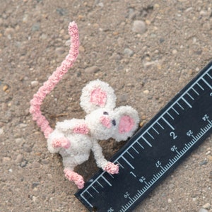 Pu&ograve; includere: Un piccolo topo bianco all'uncinetto con orecchie e coda rosa, misurato con un righello nero.