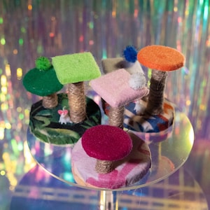 May include: A collection of miniature cat trees featuring various colored felt platforms, including green, lime, pink, orange, and magenta, supported by jute-wrapped posts on patterned circular bases, with small decorative pom-poms and a tiny white mouse figure.