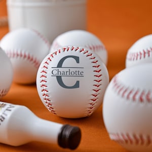 Op de afbeelding: Een witte honkbal met rode naden en de naam "Charlotte" erop gedrukt in grijs. De honkbal is omringd door andere honkballen en een fles drank.