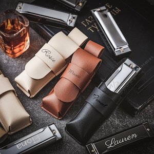 Puede incluir: Una colección de estuches de armónica personalizados en varios colores y materiales. Los estuches llevan nombres como "Rosie", "Winnie", "Jumai" y "Lauren".