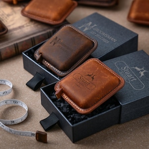 May include: Two brown leather measuring tapes with personalised engravings in black lettering. The tapes are in black boxes with black shredded paper inside. One tape says "Groomman IV" and the other says "Stuart - Est. 2019".