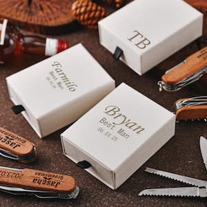 Op de afbeelding: Een assortiment gepersonaliseerde geschenkdozen en houten zakmessen. De witte dozen zijn voorzien van namen en data, en de messen hebben houten handvatten. De items zijn gerangschikt op een bruin oppervlak.