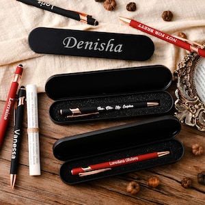 Puede incluir: Un juego de seis bolígrafos personalizados en estuches negros. Los bolígrafos son rojos con detalles dorados y tienen nombres grabados. Los nombres son "Lorraine", "Vanessa", "Denisha", "Not for Brett", "Nes. Elle. Lily. Caspian" y "Loredana Oliviero".
