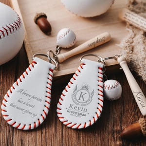 May include: White leather baseball-themed keyrings with red stitching and engraved text. One keyring reads "A brave person has a heart that never gives up." Another says "Kevin Groomsman 08.10.2024."