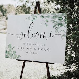 May include: A white wedding welcome sign on a wooden easel. The sign features the word "welcome" in elegant script, with the names Lillian & Doug and the date 08.10.2022. Greenery accents the top and bottom corners.