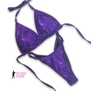 May include: A two-piece, metallic purple bikini set. The top is a triangle style with halter straps, and the bottom is a high-cut thong. The set is made of a shiny, textured fabric. The text "TOP KNOT STRONG" is visible.