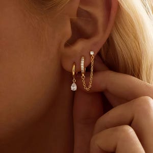 May include: Gold-toned earrings set. One earring features a teardrop-shaped clear stone, another is a small hoop, and the third is a chain connecting a small stud to a hoop earring. The earrings are worn in the ear.