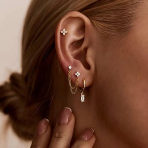 May include: An ear displaying several pieces of gold jewellery. There's a small, four-petal flower stud, a chain connecting two earrings, and a small hoop earring with a rectangular charm. All the pieces are embellished with small, clear gemstones, creating a delicate, sparkling effect.
