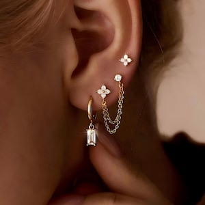 May include: Gold-toned earrings set featuring a variety of designs. Includes a hoop earring with a rectangular gem, a stud with a four-petal flower design, and a chain earring with a flower and a gem. The earrings are set against a blurred background.