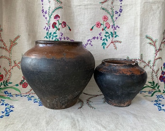 May include: Two black clay pots with brown rims, one larger than the other, sitting on a white fabric with floral embroidery.