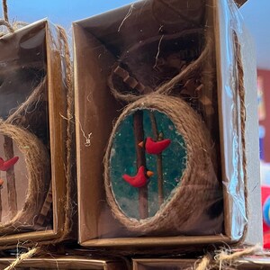 May include: Two small, clear, round ornaments with a brown twine border. Each ornament features a blue background with two red cardinals perched on branches. The ornaments are packaged in brown boxes with clear plastic lids.