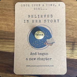 May include: A card with the text "Once upon a time, a girl... Believed in her story" and a gold charm with the word "Believe" on it. The card also has a small illustration of a book and stars. The text at the bottom of the card reads "And began a new chapter" and "www.palsinyourpocket.com".