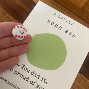 Può includere: Una spilla a forma di palla da baseball bianca con cuciture rosse e una faccia sorridente è tenuta vicino a una carta. La carta dice "A Little Home Run" e "You did it. So proud of you."
