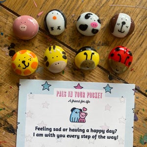 May include: Set of painted animal figurines on small rocks. The animals include a pig, penguin, cow, dog, lion, giraffe, bee, and ladybug. The rocks are arranged in two rows on a wooden surface. A card with the text "Pals in Your Pocket" and "A friend for life" is in the bottom center of the image. The card also has the text "Feeling sad or having a happy day? I am with you every step of the way!"