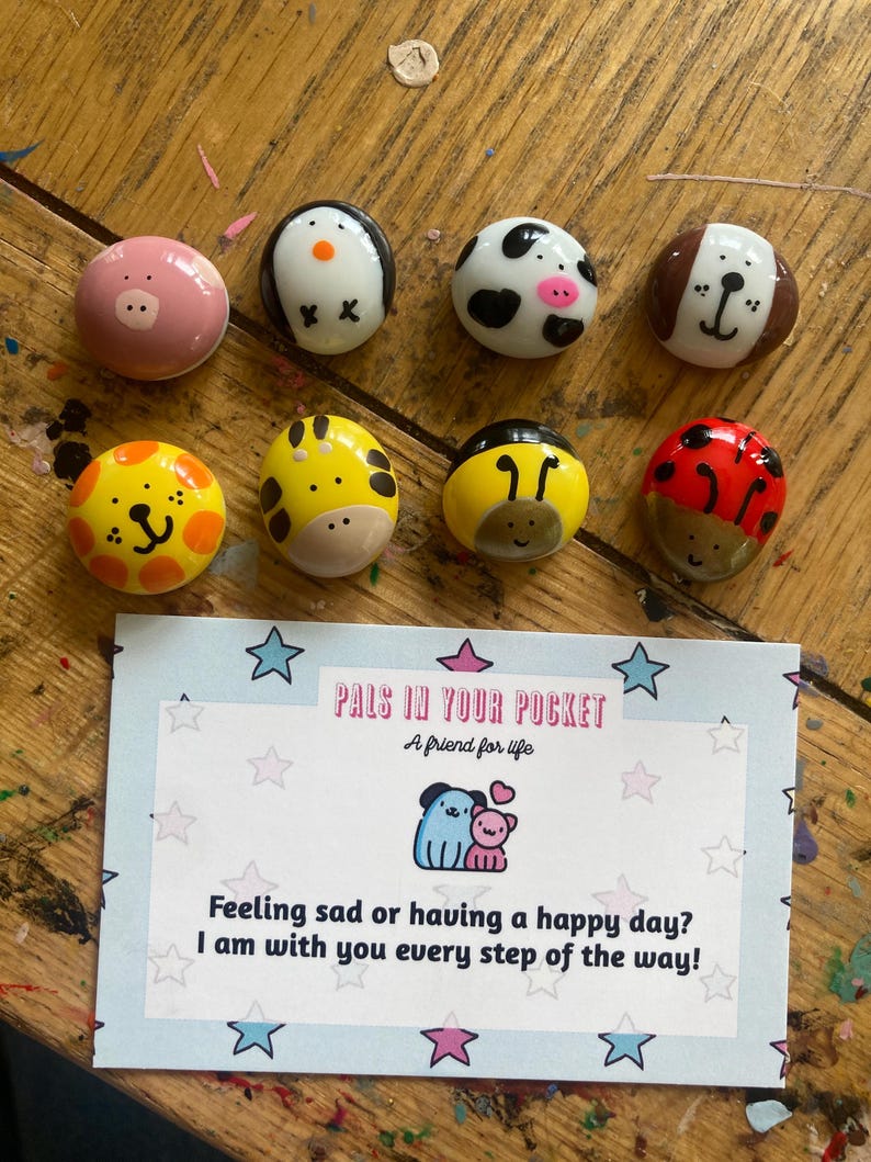 May include: Set of painted animal figurines on small rocks. The animals include a pig, penguin, cow, dog, lion, giraffe, bee, and ladybug. The rocks are arranged in two rows on a wooden surface. A card with the text "Pals in Your Pocket" and "A friend for life" is in the bottom center of the image. The card also has the text "Feeling sad or having a happy day? I am with you every step of the way!"