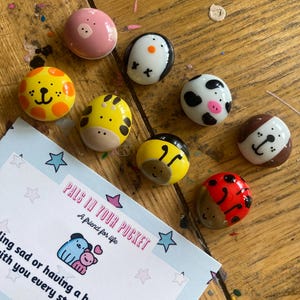 May include: A set of eight painted, round, smooth stones with different animal designs. The stones are painted in various colors, including pink, yellow, black, white, brown, red, and orange. The stones are arranged on a wooden surface with a card that reads "Pals in Your Pocket, A friend for life".