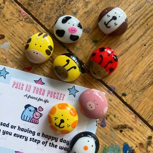 May include: A set of painted rock figurines with various animal designs, including a ladybug, a bee, a pig, a penguin, a cow, a giraffe, and a dog. The rocks are painted in bright colors and have cute faces. The rocks are labeled "Pals in Your Pocket, A friend for life".