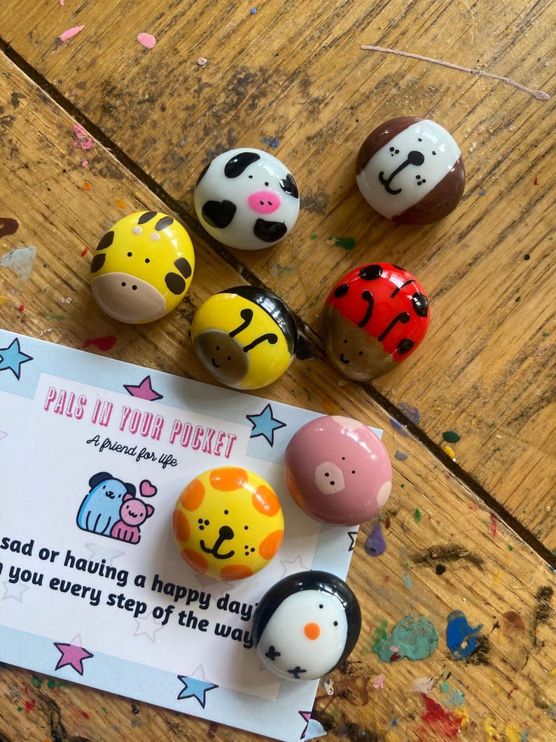 May include: A set of painted rock figurines with various animal designs, including a ladybug, a bee, a pig, a penguin, a cow, a giraffe, and a dog. The rocks are painted in bright colors and have cute faces. The rocks are labeled "Pals in Your Pocket, A friend for life".