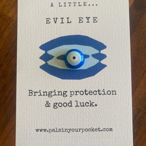 May include: A white card with blue and white text that reads "A little... Evil Eye Bringing protection & good luck."  The card also features a blue and white evil eye charm with a black pupil. The website address "www.palsinyourpocket.com" is printed at the bottom of the card.