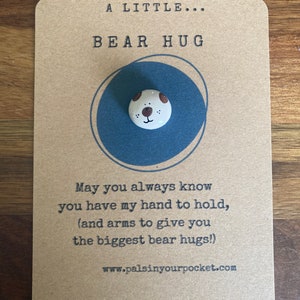 May include: A brown card with the text "A Little... Bear Hug" and a small ceramic bear face with brown eyes and a brown nose. The text below the bear face reads "May you always know you have my hand to hold, (and arms to give you the biggest bear hugs!) www.palsinyourpocket.com"