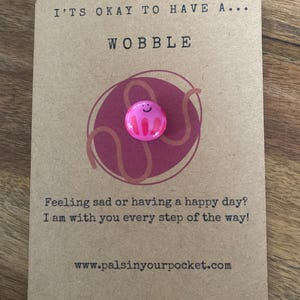 May include: A pink and red smiling jelly-like character with a white outline. The character is attached to a brown card with the text "It's okay to have a... Wobble" and "Feeling sad or having a happy day? I am with you every step of the way! www.palsinyourpocket.com".