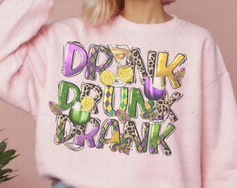 May include: A light pink sweatshirt with the words "DRINK DRUNK DRANK" in a colorful, playful design. The design includes cocktail glasses, lemons, and Mardi Gras beads. The letters have a leopard print pattern.
