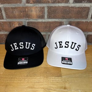 Op de afbeelding: Twee baseballpetten, één zwart en één wit, elk met het woord "JESUS" geborduurd in witte blokletters. De petten hebben een mesh achterkant en een gebogen klep. Ze liggen op een houten oppervlak.
