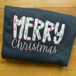 Puede incluir: Una sudadera gris oscuro con las palabras "Merry Christmas" bordadas en rosa en la parte delantera. La palabra "Merry" está compuesta por letras individuales con un fondo blanco y un patrón de pequeños árboles de Navidad.