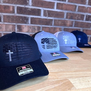 May include: Four black, gray, and blue baseball caps with an American flag and a cross design. The caps are all Richardson 112 Original Trucker adjustable.