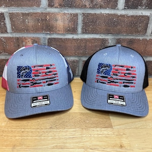 May include: Two gray trucker hats with a fishing-themed American flag design. The flag features a blue square with a fish and red and white stripes made of fishing lures. The hats have a curved brim and a Richardson 112 tag.