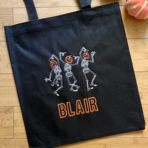 Puede incluir: Bolsa de lona negra con esqueletos bordados en blanco que llevan cabezas de calabaza y el nombre "BLAIR" en bordado naranja.