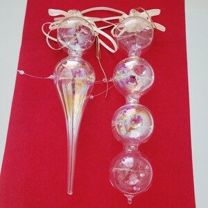 May include: Two clear glass ornaments with an iridescent finish. One ornament features a long, tapered icicle shape topped with a sphere, while the other consists of three stacked spheres. Both have cream-colored ribbons attached at the top.