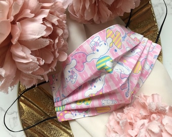 May include: Pink fabric face mask with a white background and a repeating pattern of cartoon characters. The characters are wearing pink, yellow, and blue clothing. The mask has a pleated design.