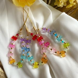 May include: Four colorful beaded phone charms with gold-colored cords. Each charm features a rainbow of small beads and flower-shaped charms. The charms are displayed on a white fabric background.