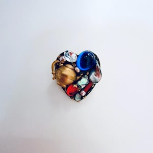 Könnte beinhalten: Eine herzförmige Brosche mit einer Ansammlung verschiedener Elemente, darunter eine kleine goldfarbene Teekanne, ein klarer facettierter Kristall, ein glatter blauer Cabochon, eine rote Perle, eine silberfarbene Tropfenform und irisierende Muschelfragmente.