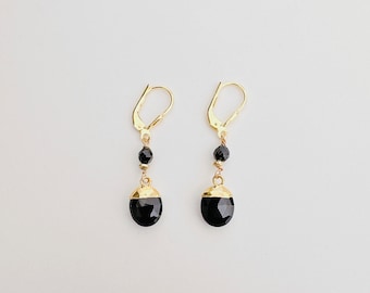 May include: A pair of gold earrings with black gemstone drops. The earrings have a simple design with a small black bead above the larger gemstone.