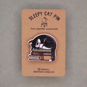 May include: Wooden pin featuring a black and white cat sleeping on a stack of books. The pin is mounted on a brown card with the text "SLEEPY CAT PIN" and "From responsibly sourced wood". The Etsy shop's Instagram handle and website are also displayed.