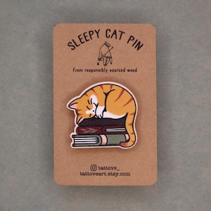 May include: A wooden pin featuring a sleeping orange tabby cat atop a stack of books. The pin is on a brown card with the text "SLEEPY CAT PIN" and "from responsibly sourced wood". The Etsy shop's Instagram handle and website are also visible.