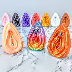 May include: A collection of 10 colorful ceramic sculptures in various shapes and sizes. The sculptures are arranged in a semi-circle on a white marble surface. The sculptures are all abstract and feature a variety of colors, including pink, purple, blue, green, orange, and yellow.
